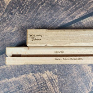 Può includere: Due binari in legno chiaro per appendere poster con un design scanalato. Il binario superiore riporta il marchio Haftowany Zakątek. Il binario inferiore mostra il testo 40cm/16in e Made in Poland, Cieszyn 2024, indicando la lunghezza.
