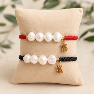 May include: Two bracelets featuring four white baroque pearls each, separated by small gold-tone beads. One bracelet has a red woven cord, the other a black woven cord. Both include a small gold-tone bear-shaped charm hanging from the side.