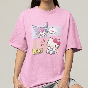 May include: Pink t-shirt with a graphic print of Hello Kitty and a character with a skull on its head. The text "Doki" is printed on the shirt.