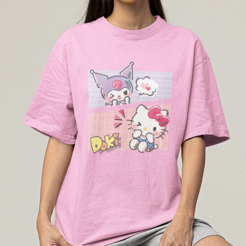 May include: Pink t-shirt with a graphic print of Hello Kitty and a character with a skull on its head. The text "Doki" is printed on the shirt.