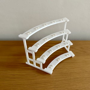 May include: White three-tiered curved shelf made of LEGO bricks. The shelf is designed to hold small items, such as jewelry or other trinkets.
