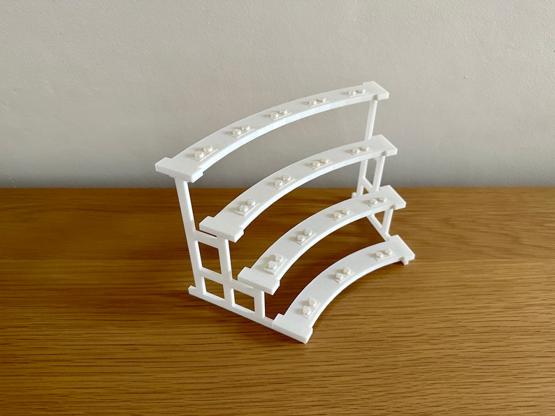 May include: White three-tiered curved shelf made of LEGO bricks. The shelf is designed to hold small items, such as jewelry or other trinkets.