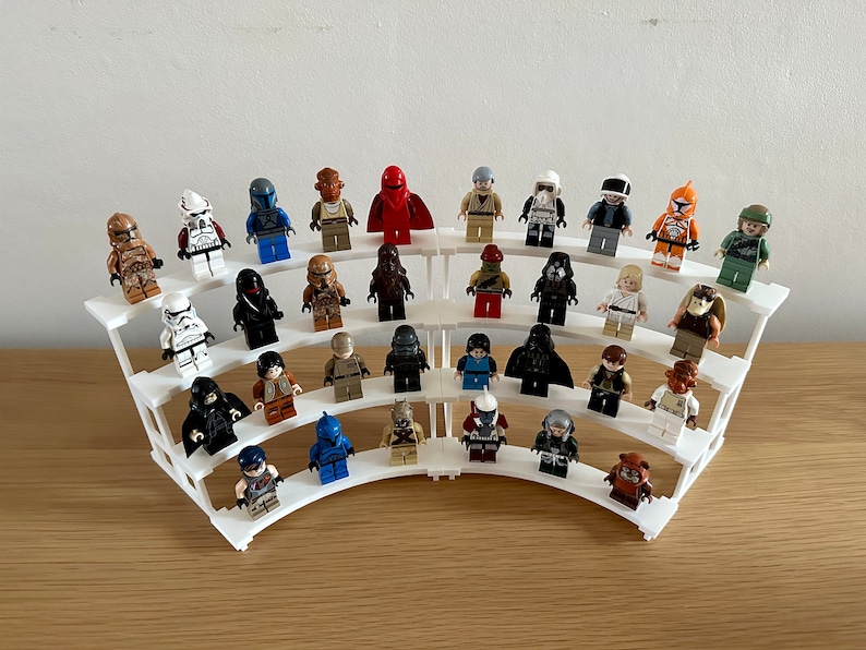 May include: A white plastic display stand with multiple tiers holds a collection of Star Wars minifigures. The figures are arranged in a semi-circle and include characters like Darth Vader, Luke Skywalker, and Stormtroopers.