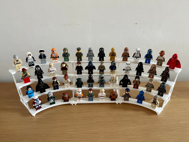 May include: A collection of 36 Star Wars LEGO minifigures displayed on a white tiered shelf. The minifigures are arranged in rows and include characters such as Darth Vader, Yoda, and Boba Fett.