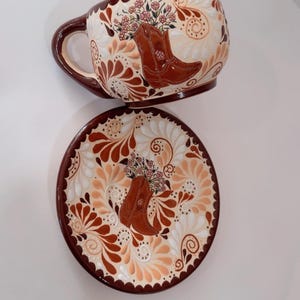 May include: A ceramic cup and saucer set with a Western theme. The cup and saucer are decorated with a brown cowboy boot filled with flowers. The background features a floral pattern in cream, brown, and peach tones.