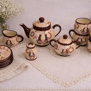 May include: A ceramic tea set with a brown and cream floral design, including a teapot, sugar bowl, creamer, cups, and saucers. Each piece features a horseshoe motif. The set is arranged on a white tablecloth with lace trim.