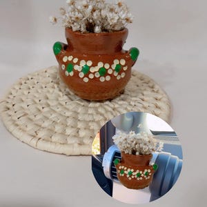 May include: A small terracotta pot with green accents and white dots, filled with dried white flowers. The pot is sitting on a woven straw mat.