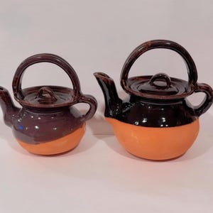 May include: Two ceramic teapots. The teapots have a dark brown handle, lid, and spout. The lower half of the teapots are orange, and the upper half is a dark purple. The teapots are side by side.