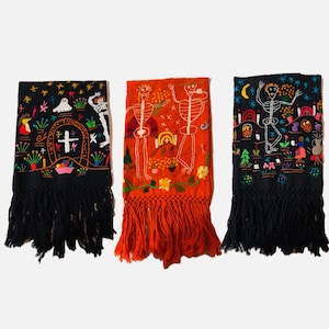 May include: Three embroidered Day of the Dead scarves. One is orange, one is black, and one is black. Each scarf features a detailed design of skeletons, flowers, and other festive elements. The scarves have a fringe at the bottom.