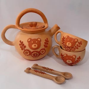 May include: A terracotta tea set featuring a teapot, two teacups, and two spoons. The teapot and cups are decorated with a bear face and floral designs. The spoons have bear head details. The set is made of ceramic.