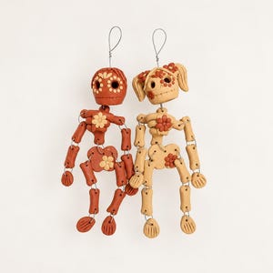 May include: Two articulated skeleton ornaments made of clay, one in dark terracotta and one in light beige. Each figure features floral details on the head, chest, and pelvis, with limbs connected by silver wire loops for hanging display.