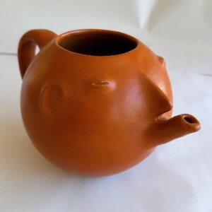 May include: A terracotta teapot with a whimsical face and a spout shaped like a nose. The teapot is a rounded shape and has a handle on the side.