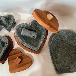 May include: Heart-shaped mortar and pestle sets. The mortar is made of gray stone and sits on a wooden base. The pestle is also gray stone. The wooden lids have a small wooden square on top. The set is designed for grinding spices and herbs.