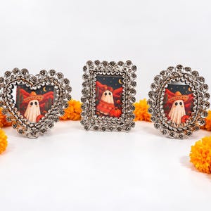May include: Three silver-toned metal frames with intricate floral-like edges, each containing a print of a ghost wearing a sombrero in a desert landscape with cacti and pumpkins, surrounded by bright orange marigold flowers.