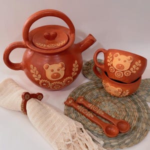 May include: A terracotta-colored tea set featuring a teapot, two cups, spoons, and napkin rings, all adorned with bear designs. The set includes a woven placemat and a fringed napkin.