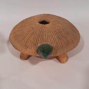 May include: A ceramic turtle-shaped decorative item. The turtle has a brown, textured shell with a central opening. The head is green and the feet are tan. The item is designed to be a decorative piece.