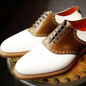 Puede incluir: Dos pares de zapatos de montar de cuero blanco y marrón con cordones. Los zapatos tienen una parte superior de lona blanca y una puntera de cuero marrón.