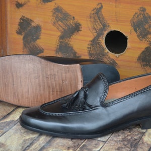 K&ouml;nnte beinhalten: Schwarze Leder-Tasssel-Slipper mit brauner Ledersohle. Die Slipper haben ein gen&auml;htes Detail am Rand des Schuhs und einen Quasten an der Oberseite.
