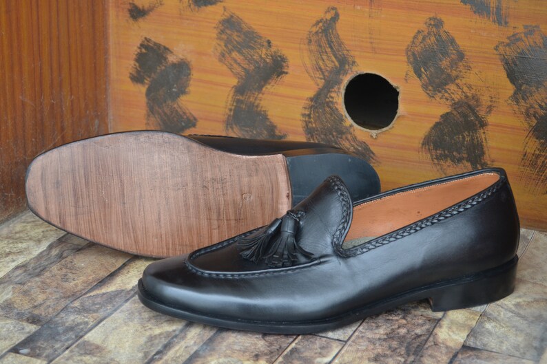 K&ouml;nnte beinhalten: Schwarze Leder-Tasssel-Slipper mit brauner Ledersohle. Die Slipper haben ein gen&auml;htes Detail am Rand des Schuhs und einen Quasten an der Oberseite.