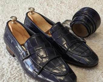 May include: Two dark blue leather loafers with a crocodile pattern and a buckle closure. The loafers are on a light beige surface.