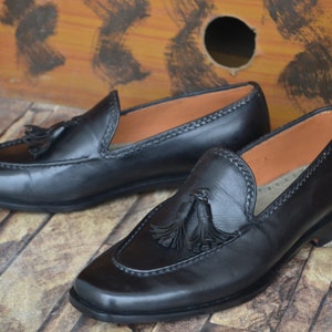K&ouml;nnte beinhalten: Ein Paar schwarze Leder-Tasssel-Slipper mit einem Steppdetail um die &Ouml;ffnung. Die Slipper haben eine abgerundete Spitze und einen niedrigen Absatz.