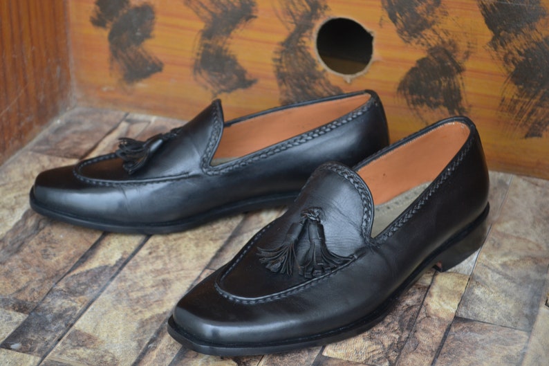 K&ouml;nnte beinhalten: Ein Paar schwarze Leder-Tasssel-Slipper mit einem Steppdetail um die &Ouml;ffnung. Die Slipper haben eine abgerundete Spitze und einen niedrigen Absatz.