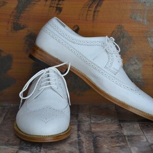 May include: A pair of white leather brogue shoes with brown leather soles. The shoes have a lace-up closure and a wingtip design.