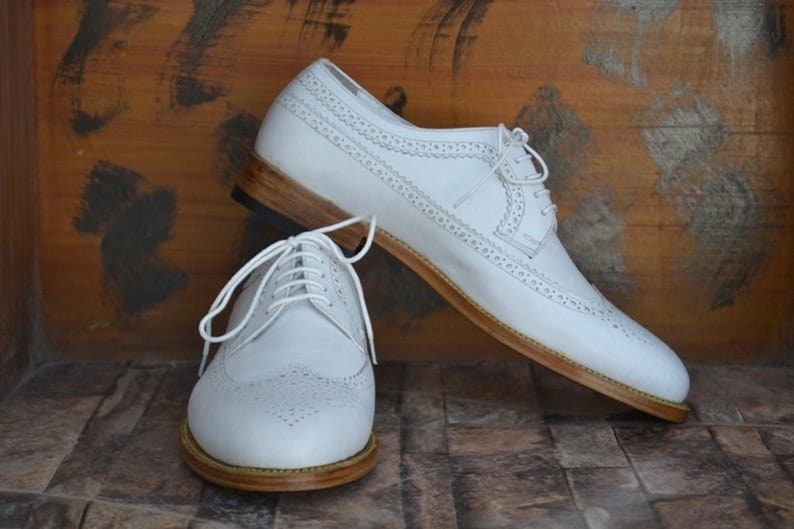 May include: A pair of white leather brogue shoes with brown leather soles. The shoes have a lace-up closure and a wingtip design.