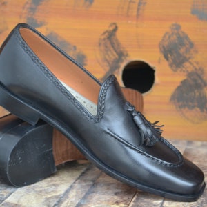 K&ouml;nnte beinhalten: Schwarze Leder-Tasssel-Slipper mit einem gestickten Detail am Rand. Die Slipper liegen auf einer Holzoberfl&auml;che.