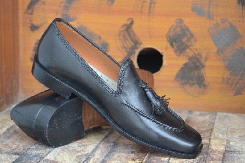 K&ouml;nnte beinhalten: Schwarze Leder-Tasssel-Slipper mit einem gestickten Detail am Rand. Die Slipper liegen auf einer Holzoberfl&auml;che.