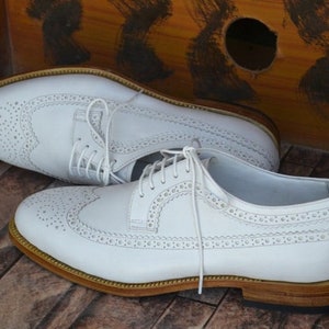 May include: A pair of white leather brogue shoes with a lace-up closure and a brown leather sole.