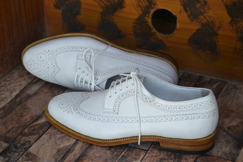 May include: A pair of white leather brogue shoes with a lace-up closure and a brown leather sole.