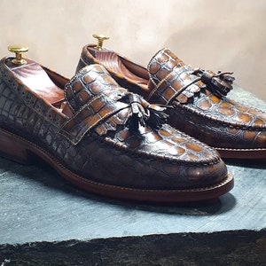 May include: A pair of brown leather loafers with a crocodile pattern and tassel accents. The loafers have a brown leather sole and a brown leather upper.