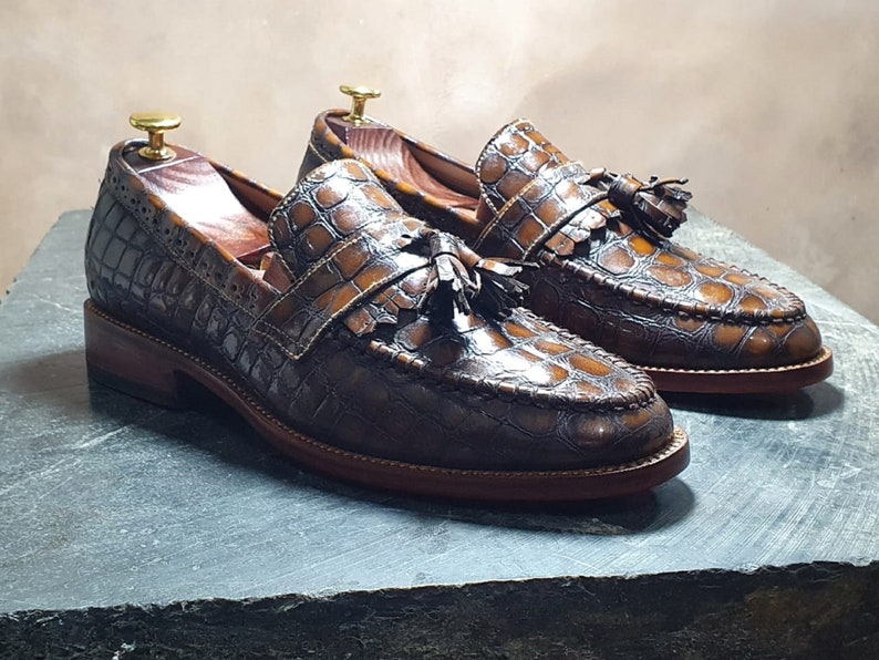 May include: A pair of brown leather loafers with a crocodile pattern and tassel accents. The loafers have a brown leather sole and a brown leather upper.