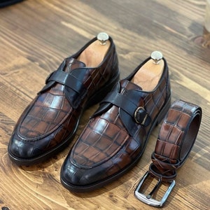 Pode incluir: Um par de sapatos de couro marrom com padrão de crocodilo e fivelas pretas. Os sapatos têm bico fino e salto baixo. Um cinto de couro marrom combinando com padrão de crocodilo também está incluído.