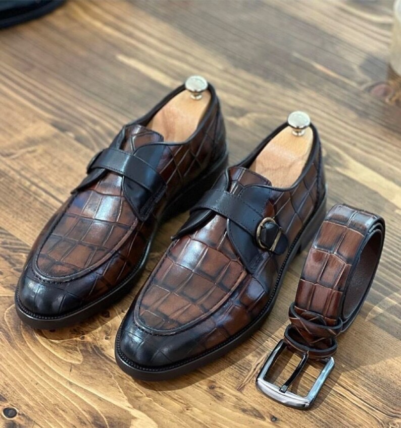 May include: A pair of brown leather shoes with a crocodile pattern and black buckles. The shoes have a pointed toe and a low heel. A matching brown leather belt with a crocodile pattern is also included.