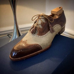 Op de afbeelding: Bruine en beige leren nette schoenen met kwastjes veters. De schoenen hebben een wingtip ontwerp en een spitse neus.
