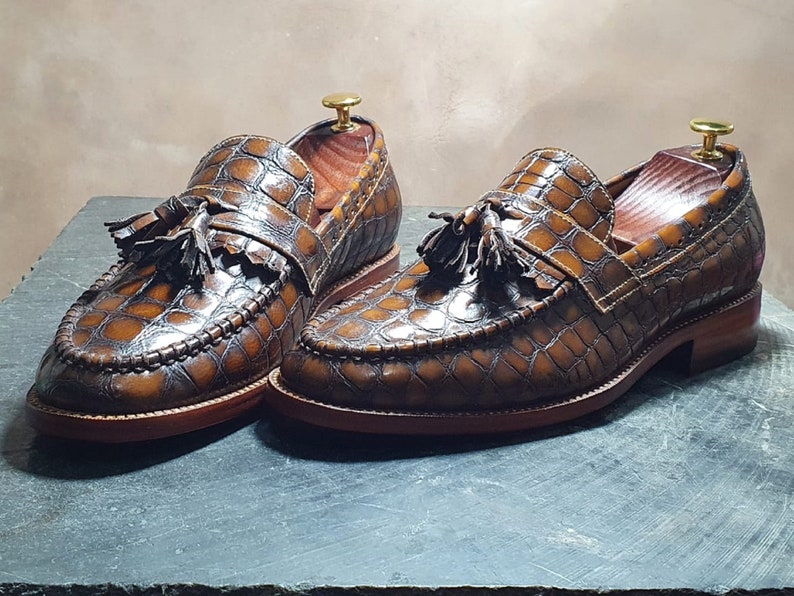 May include: A pair of brown leather loafers with a crocodile pattern and tassel accents. The loafers have a brown leather sole and a stitched edge.