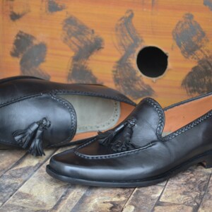K&ouml;nnte beinhalten: Ein Paar schwarze Leder-Tasssel-Slipper mit einem gestickten Detail am Rand. Die Slipper liegen auf einer holzartigen Oberfl&auml;che.