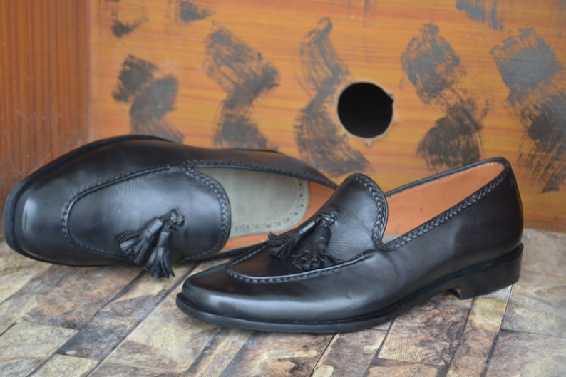 K&ouml;nnte beinhalten: Ein Paar schwarze Leder-Tasssel-Slipper mit einem gestickten Detail am Rand. Die Slipper liegen auf einer holzartigen Oberfl&auml;che.