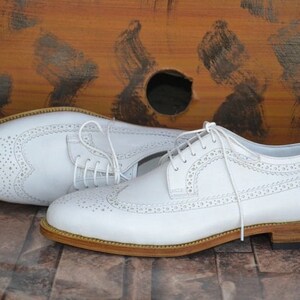 May include: A pair of white leather brogue shoes with a lace-up closure and a brown leather sole.