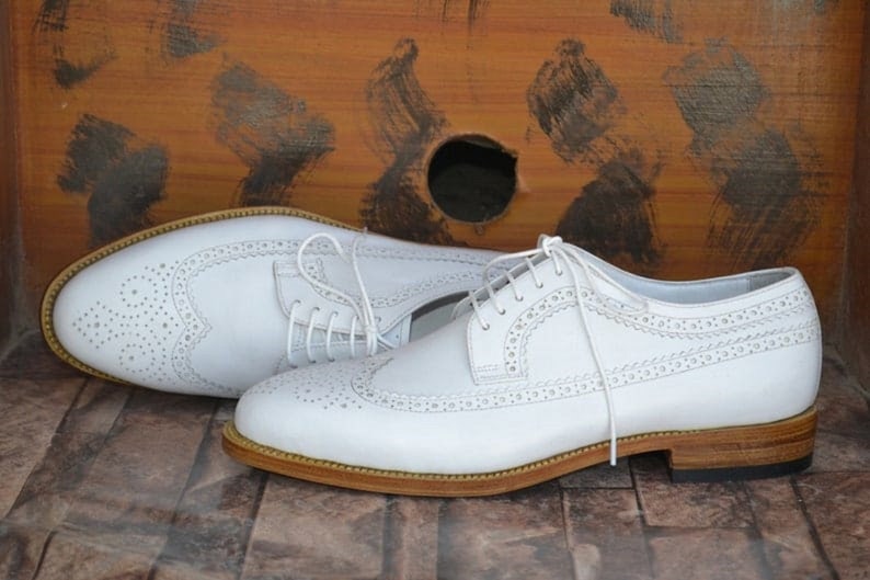 May include: A pair of white leather brogue shoes with a lace-up closure and a brown leather sole.