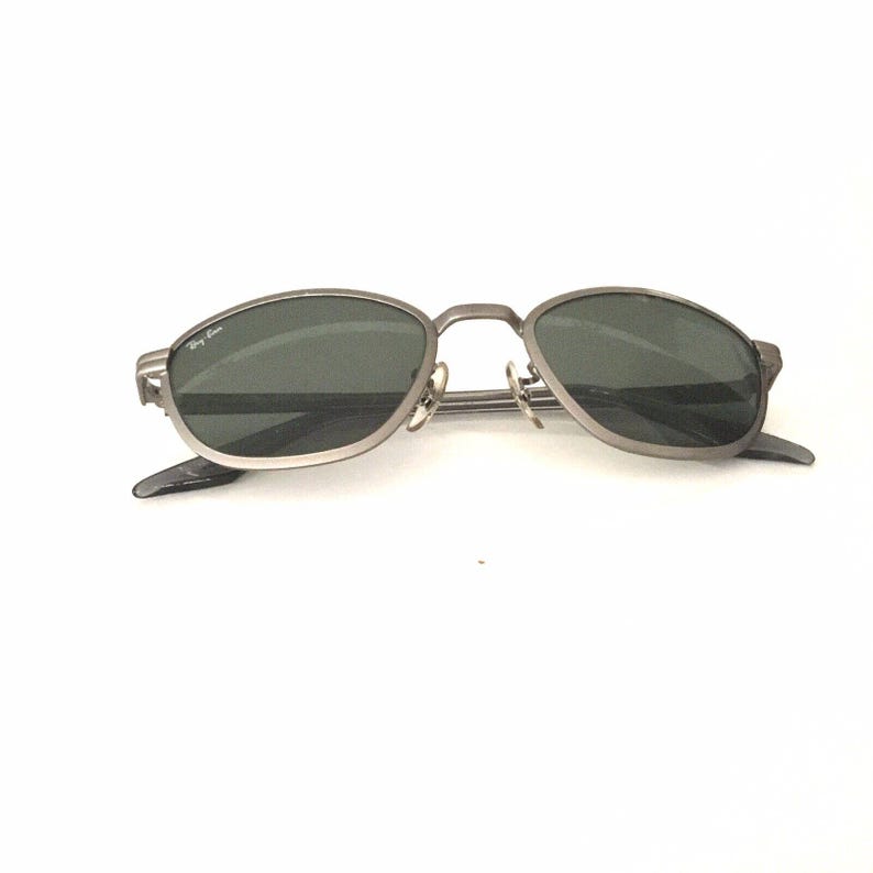 May include: A pair of silver-toned sunglasses with dark gray lenses. The sunglasses have a thin metal frame and a Ray-Ban logo on the left temple.
