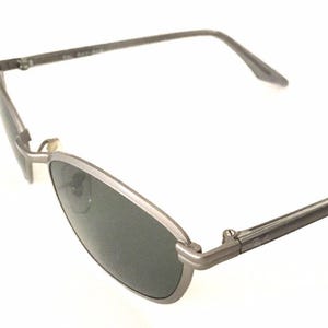 May include: Silver-toned metal sunglasses with dark gray lenses. The sunglasses have a thin frame and a slightly curved shape.