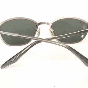 May include: Silver-toned oval sunglasses with dark gray lenses. The sunglasses are shown from the back, with the arms crossed.