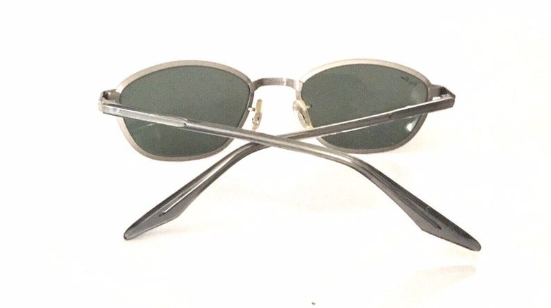 May include: Silver-toned oval sunglasses with dark gray lenses. The sunglasses are shown from the back, with the arms crossed.