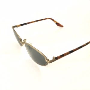 Puede incluir: Gafas de sol ovaladas con montura de metal dorado y lentes oscuras. Las patillas presentan un patr&oacute;n de carey en tonos marrones y negros. Las gafas de sol est&aacute;n anguladas, mostrando su dise&ntilde;o.