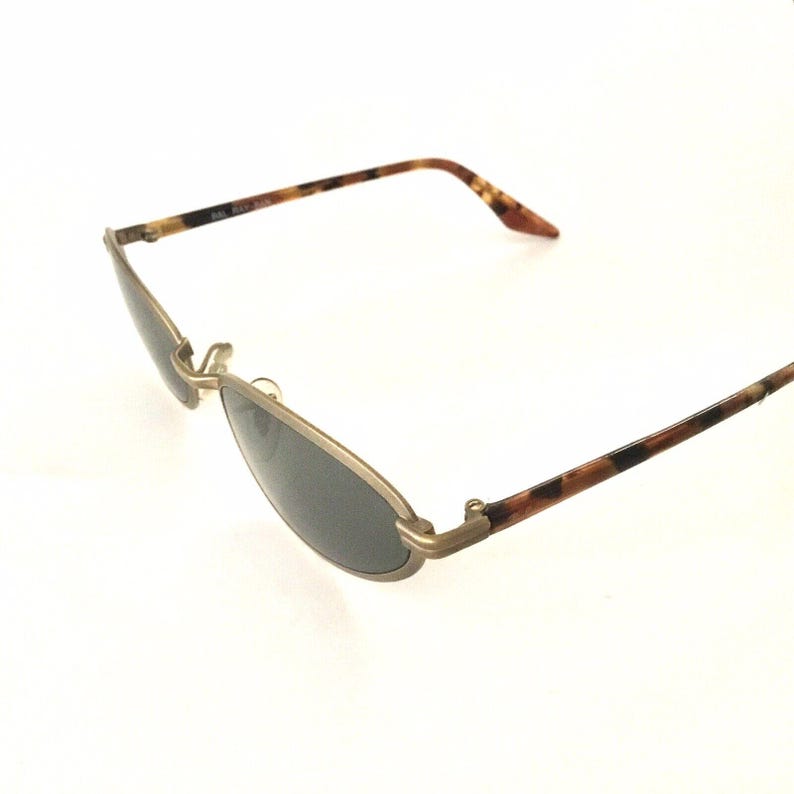 Puede incluir: Gafas de sol ovaladas con montura de metal dorado y lentes oscuras. Las patillas presentan un patr&oacute;n de carey en tonos marrones y negros. Las gafas de sol est&aacute;n anguladas, mostrando su dise&ntilde;o.