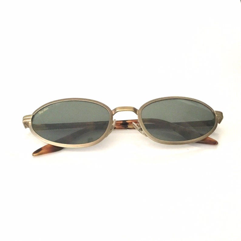 Puede incluir: Gafas de sol ovaladas con montura de metal dorado y lentes oscuras. Las patillas presentan un dise&ntilde;o de carey. Las gafas se muestran sobre un fondo blanco.