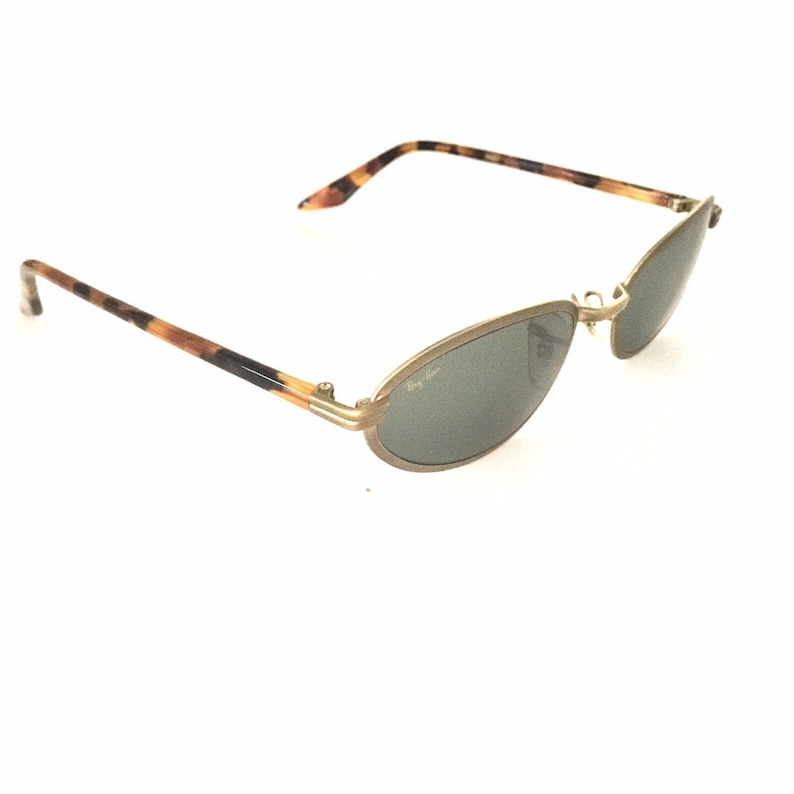 Puede incluir: Gafas de sol Ray-Ban vintage con lentes ovaladas y montura de metal dorado. Las patillas presentan un patr&oacute;n de carey. El logotipo de Ray-Ban es visible en la lente. Un accesorio de gafas cl&aacute;sico.